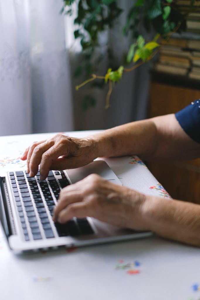 Elderly person typing on a laptop indoors, showing technology usage in daily life.